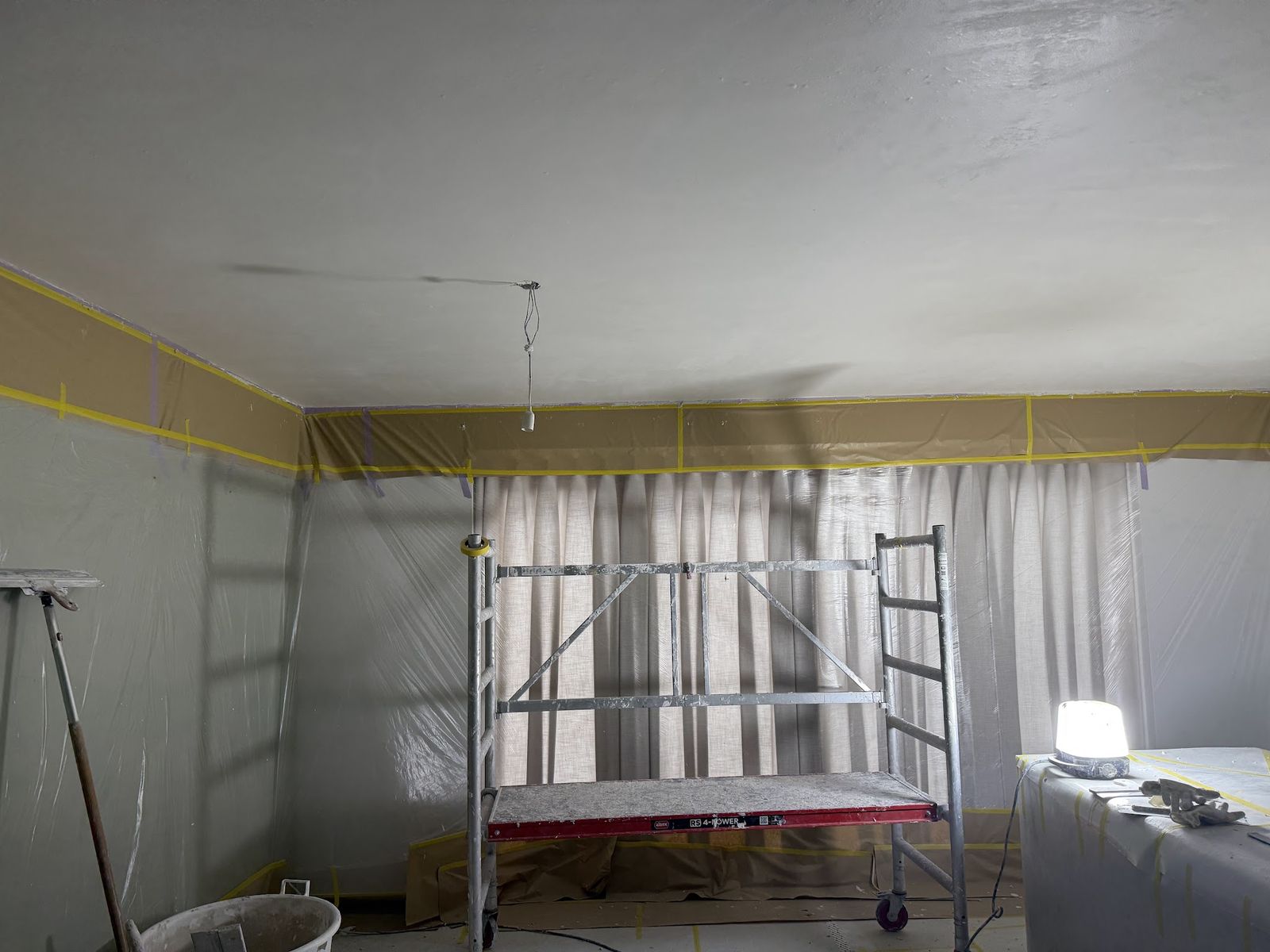 Een plafond wordt gestuct vanaf een rolsteiger, wanden en gordijnen zijn met folie afgedekt